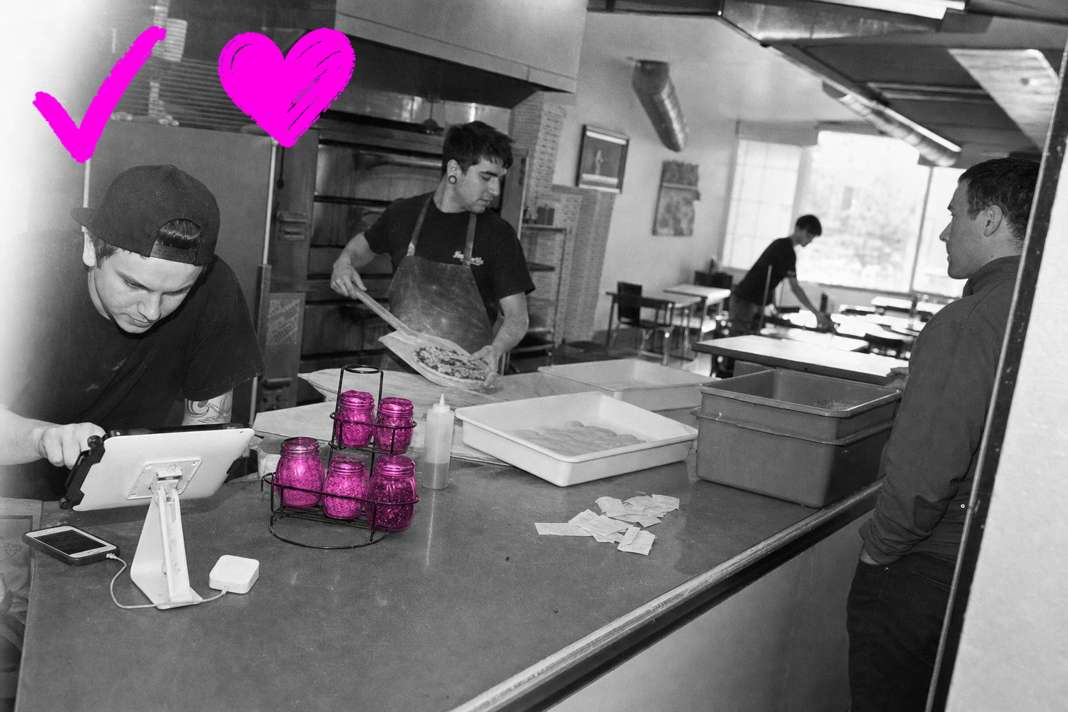 Wide black-and-white photo inside a busy small pizzeria: a server checks a frozen order tablet at the register, a cook slides a pizza onto a peel behind the counter, a customer waits by the door and someone buses a table by the window, with a rack of counter jars washed in hot magenta.
