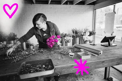 Black-and-white point-and-shoot photograph of a young tattooed florist mid-arrangement at a cluttered worktable inside a small flower shop, 2015; a loose bunch of ranunculus blooms in her hand glows with a translucent hot-magenta wash, the only color in the frame.