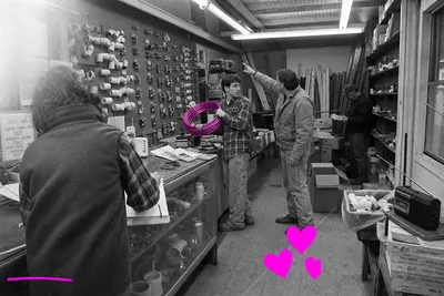 Wide black-and-white 35mm snapshot of a cluttered 1997 plumbing supply shop interior: a woman writes on a clipboard at the counter in the foreground, a young worker mid-frame hoists a coil of copper pipe — washed in a translucent hot-magenta highlight — toward a customer pointing at the pegboard wall, while an older man browses shelves alone in the background.