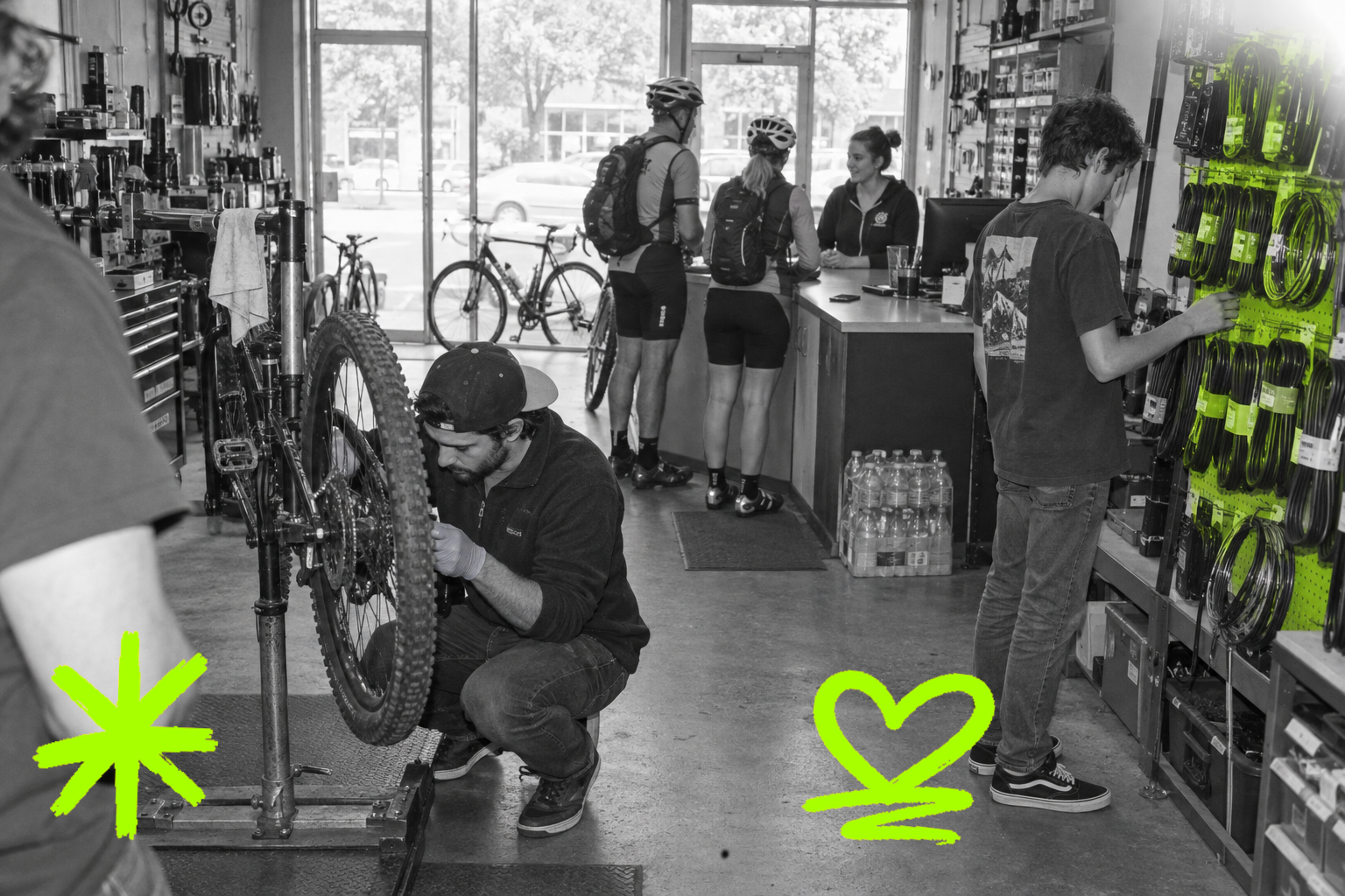 Wide black-and-white point-and-shoot interior of a bike repair shop in 2014: a mechanic in a snapback crouches over an upturned mountain bike mid-left, a teenager browses a pegboard of inner tubes mid-right — those tubes glowing in a translucent acid-green wash — while two helmeted cyclists talk to a counter attendant in the bright background and a fifth person's shoulder enters the left edge of frame.